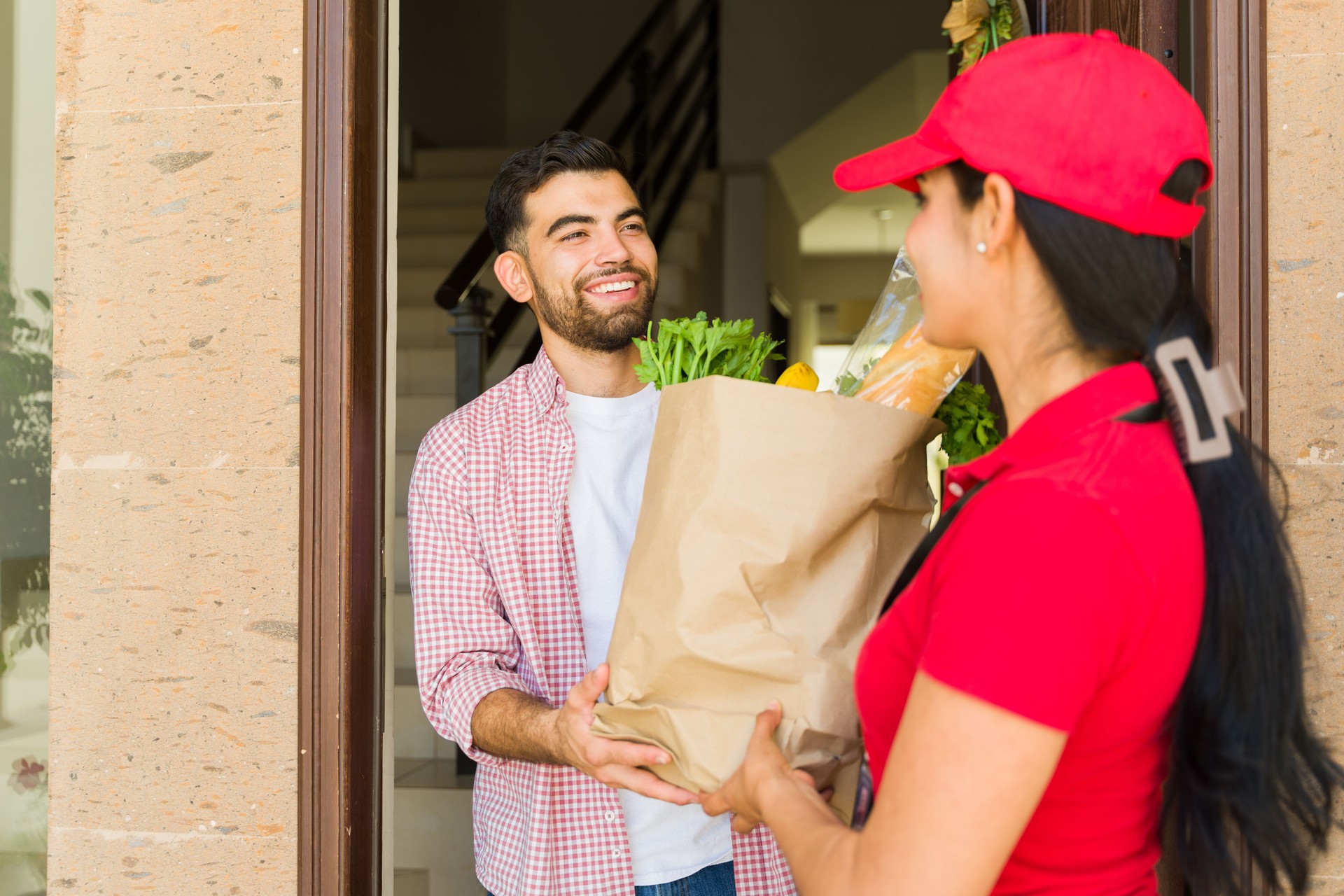 Delivery driver in a red cap handing a grocery bag to a smiling customer at the front door.