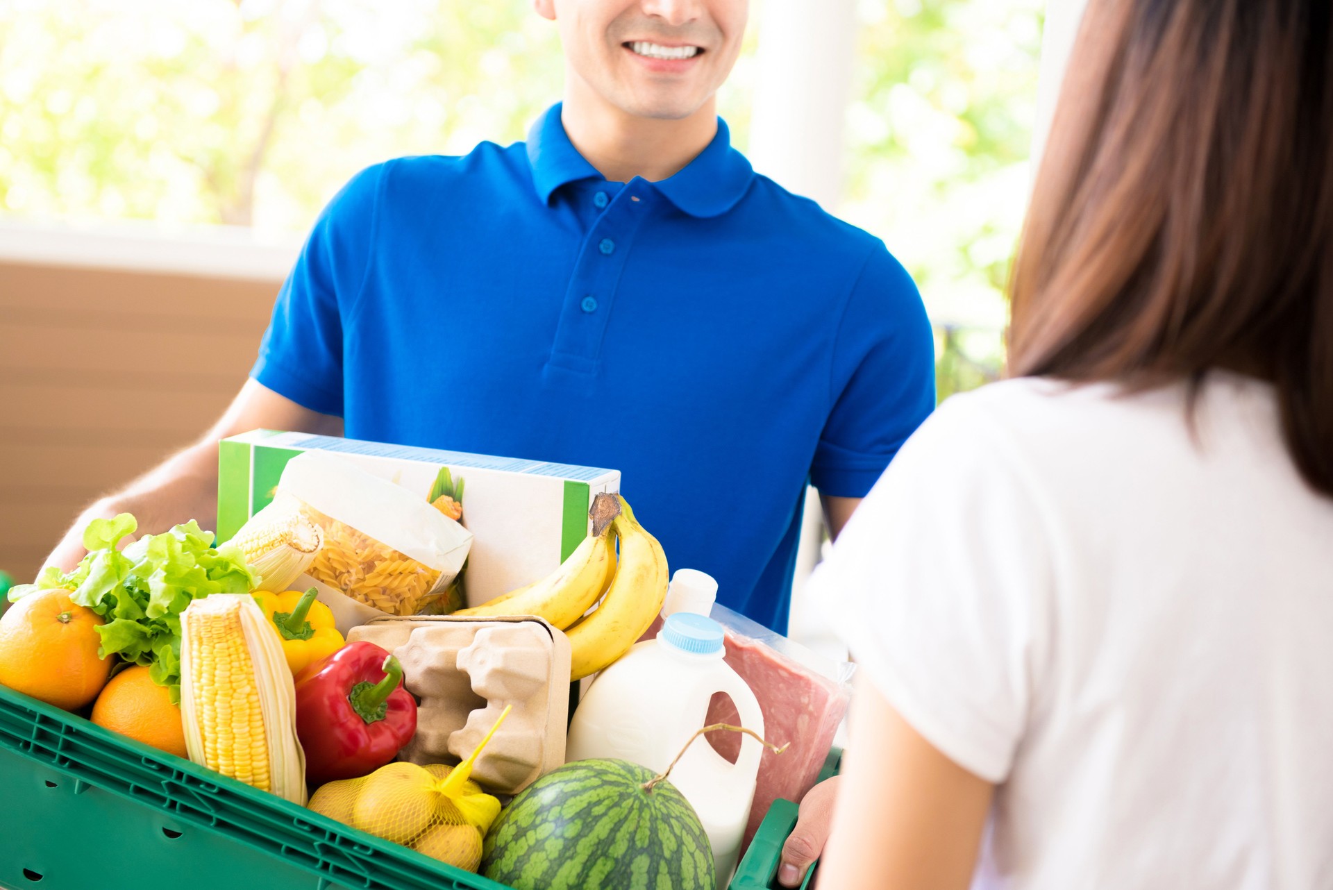 Delivery driver in a blue polo shirt handing a box of fresh groceries to a customer at the door.